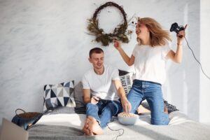 Um Jovem Casal Sentado Em Uma Cama Em Casa, Jogando Videogame Juntos Enquanto Saboreiam Pipoca, Criando Uma Atmosfera Aconchegante E Divertida Para O Final De Semana.
