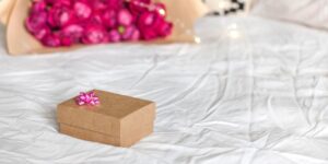 Uma Caixa De Presente Lindamente Embrulhada Com Um Buquê De Rosas, Repousando Em Uma Mesa De Cabeceira Ao Lado De Uma Cama, Com Um Fundo Desfocado Suave.