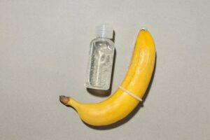 Uma Banana Usando Um Preservativo Ao Lado De Uma Garrafa De Lubrificante, Destacando O Tema Da Saúde E Segurança Sexual.