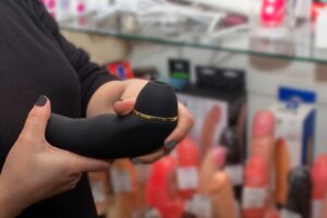 Um Funcionário De Uma Sex Shop Apresentando Uma Variedade De Brinquedos Sexuais Coloridos A Um Cliente Em Um Ambiente Discreto.