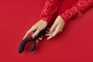 Um Par De Mãos Femininas Com Esmalte Rosa, Vestindo Mangas Vermelhas, Segurando Um Vibrador Preto Contra Um Fundo Vermelho.